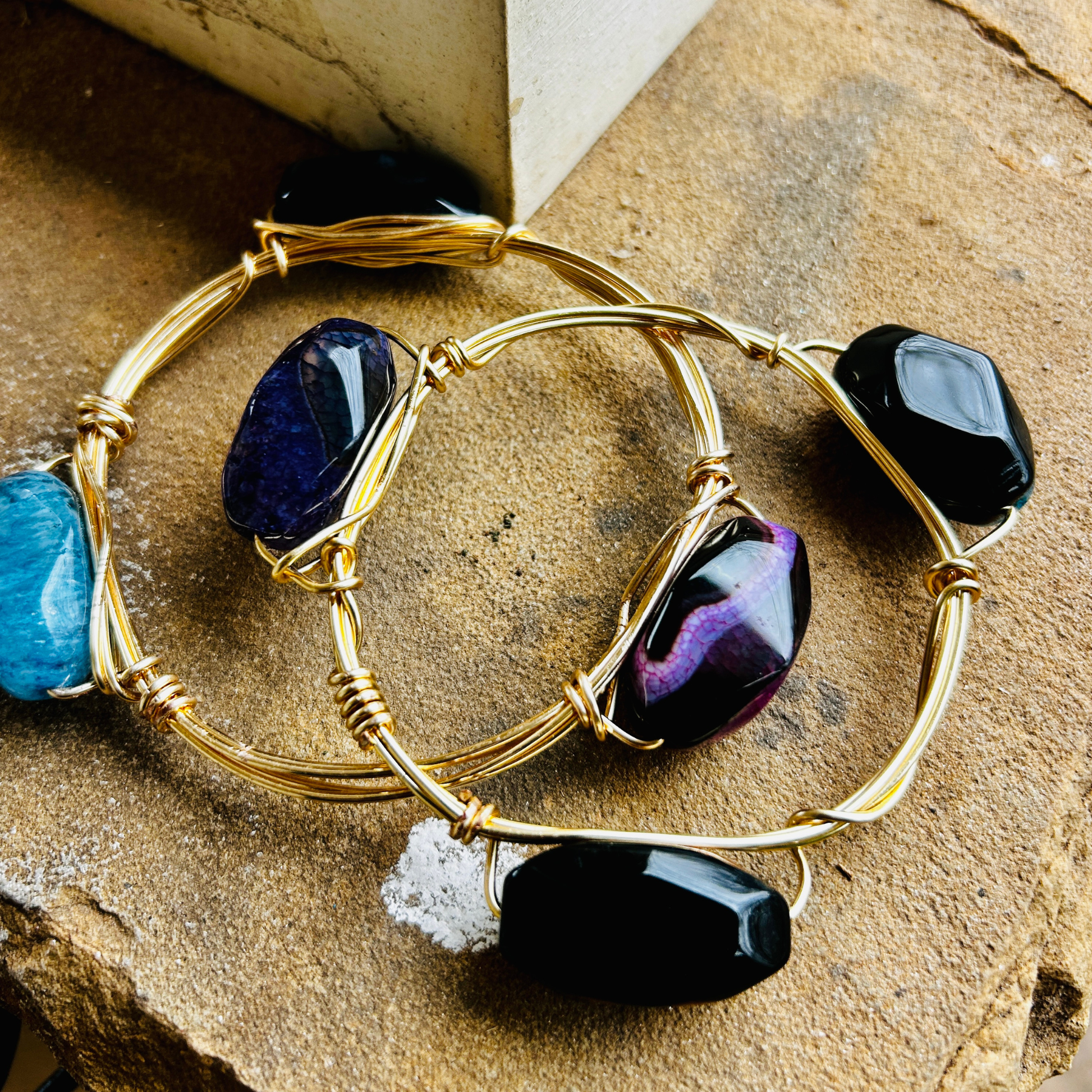 “Handcrafted gold wire-wrapped gemstone bangles featuring black onyx, purple druzy, and blue agate stones on a rustic stone background”