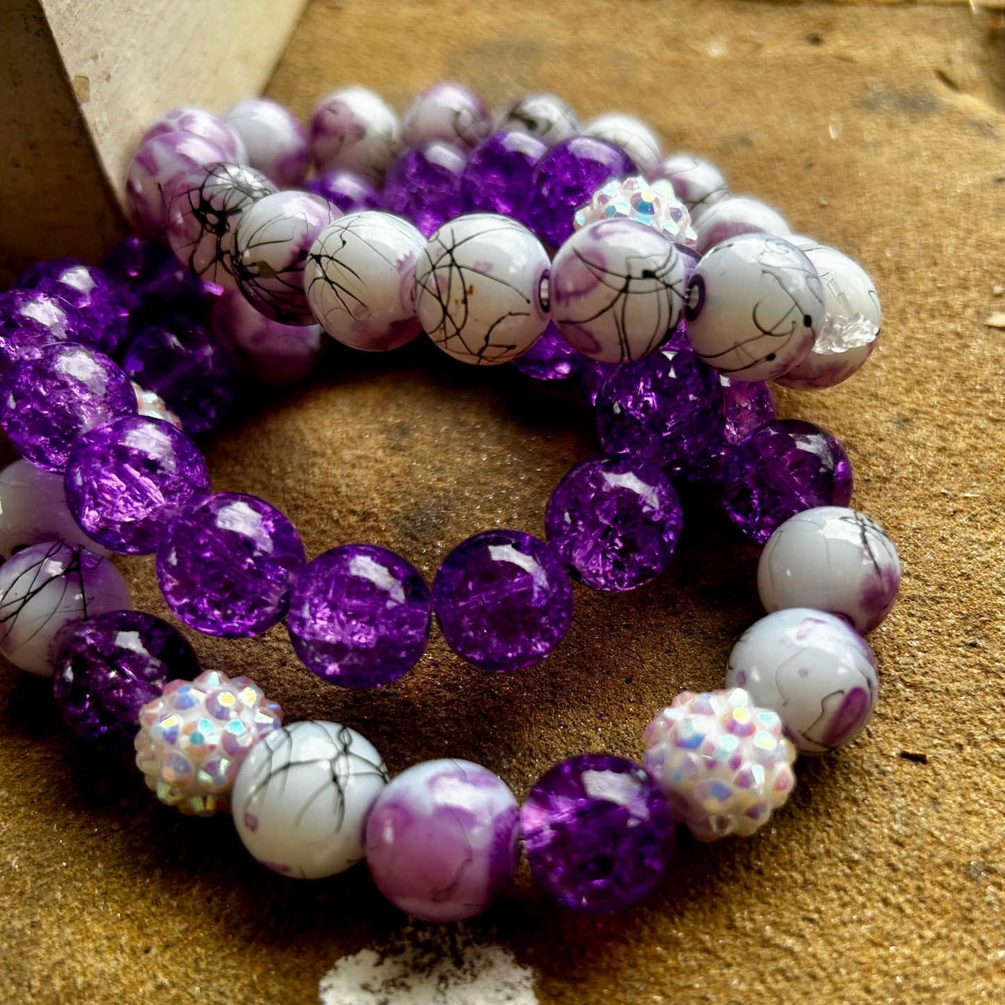 A vibrant handmade bracelet set featuring glossy purple crackle beads and white marble swirl beads with iridescent rhinestone spacers, displayed on a textured tan stone surface.